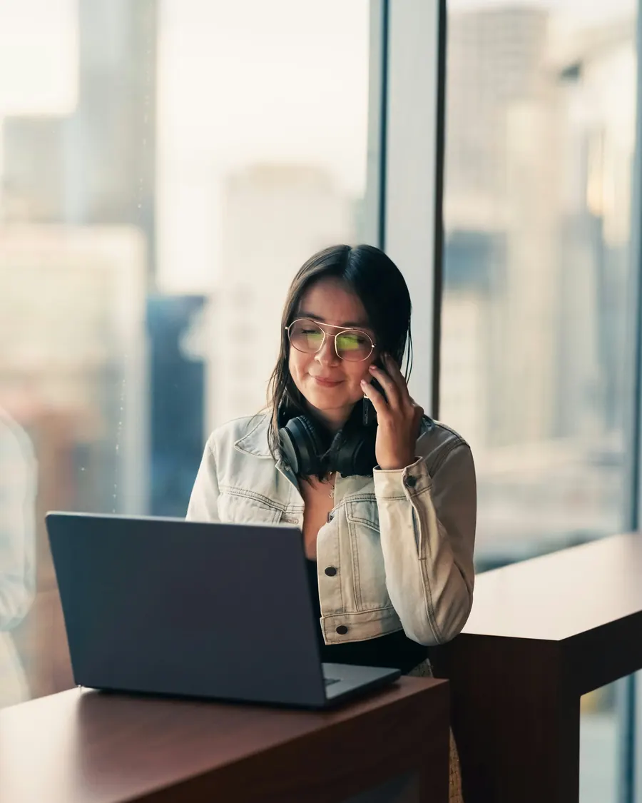 Young woman on a phone call at her laptop by a city window